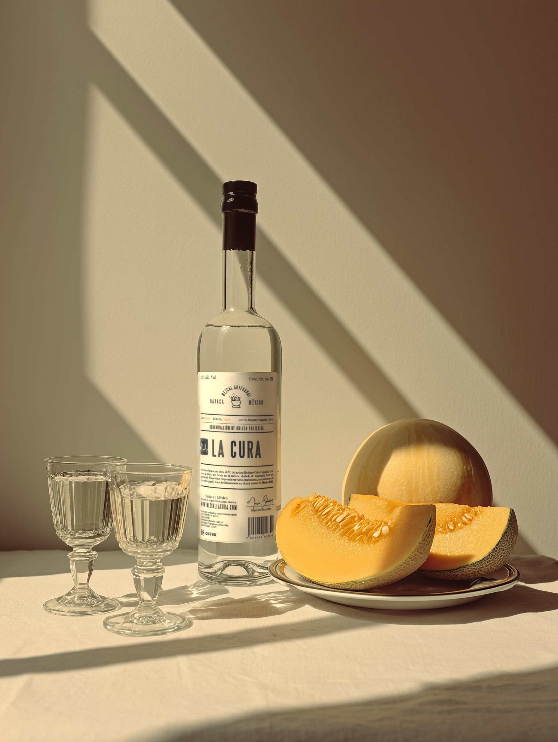 Mezcal LA CURA Espadín 45% junto a melón fresco y copas sobre mesa iluminada. Mezcal artesanal de Miahuatlán con perfil sensorial extraordinario.