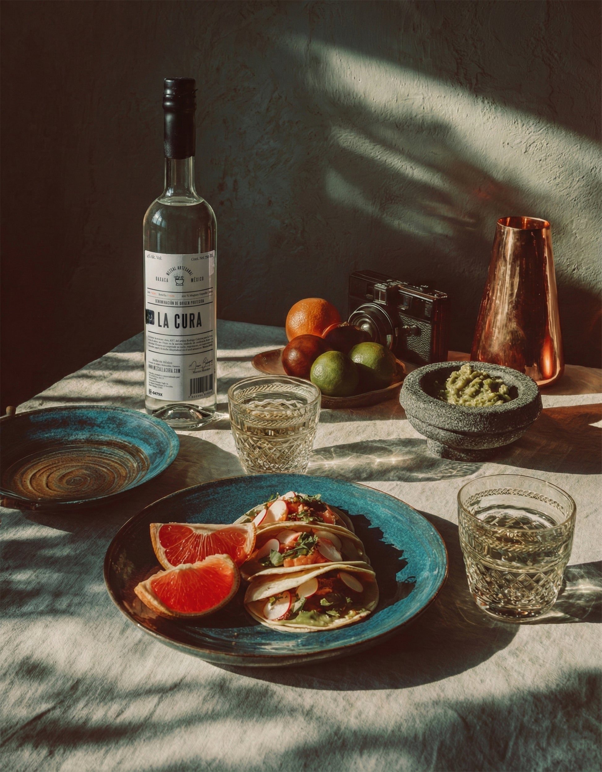 Botella de Mezcal LA CURA con comida mexicana en mesa artesanal iluminada por luz natural.